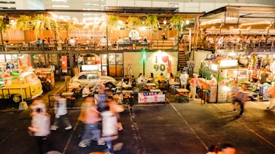 An outdoor food market is bustling with people walking around and others sitting at tables enjoying meals. The scene includes vibrant food stalls, colorful lighting, and greenery hanging from above. The environment looks lively and social, with a mix of retro and modern decor.