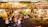 An outdoor food market is bustling with people walking around and others sitting at tables enjoying meals. The scene includes vibrant food stalls, colorful lighting, and greenery hanging from above. The environment looks lively and social, with a mix of retro and modern decor.