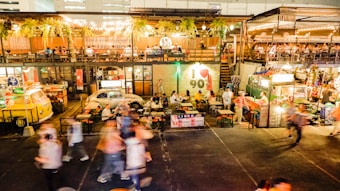An outdoor food market is bustling with people walking around and others sitting at tables enjoying meals. The scene includes vibrant food stalls, colorful lighting, and greenery hanging from above. The environment looks lively and social, with a mix of retro and modern decor.