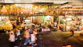 An outdoor food market is bustling with people walking around and others sitting at tables enjoying meals. The scene includes vibrant food stalls, colorful lighting, and greenery hanging from above. The environment looks lively and social, with a mix of retro and modern decor.