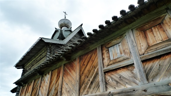 A wooden building features intricate craftsmanship with interlocking wooden beams and an ornate roof structure. The roof is topped with an onion-shaped dome, commonly found in traditional Eastern European architecture. The wood shows variations in color and texture, indicative of natural aging.