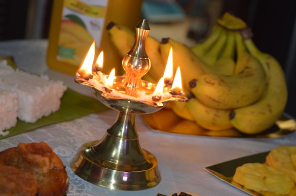 A lit brass oil lamp casting a warm glow, surrounded by a variety of items including ripe bananas and a bottle with a yellow label. There are other food items on banana leaves, suggesting a festive or ritualistic setting.