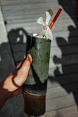 A hand holding a bamboo cup with an orange straw and a white orchid flower garnished on top. Shadows of the hand and cup are cast on a wooden surface in the background.