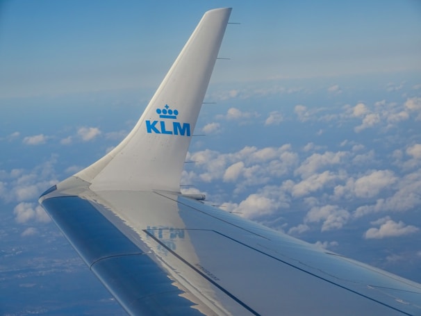 An airplane wing extends into a clear sky with scattered clouds below, featuring the KLM logo on the wingtip against a backdrop of vibrant blue.