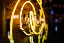 Warm yellow neon light tubes arranged artistically against a wooden background, reflecting in the glass with bokeh light effects in the distance.