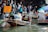 A busy floating market scene where several people are in boats on the water. In the foreground, a person wearing a helmet and holding a smartphone is seated in a boat with a paddle. Another person in a neighboring boat wears a hat. There is a sign in the background offering 'Satay Chicken Pork.' The market is filled with people and a variety of goods being traded.