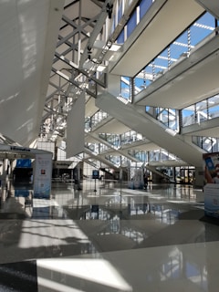A spacious, modern architectural interior featuring large windows with multiple beams of sunlight streaming through. The design includes geometric patterns with high ceilings and intersecting beams. There are informational signages and posters on the polished floor beneath a well-lit environment.