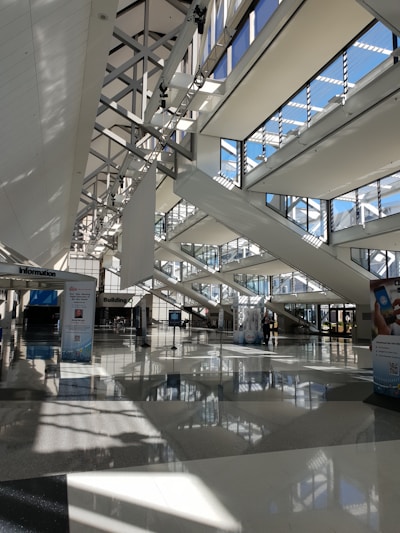 A spacious, modern architectural interior featuring large windows with multiple beams of sunlight streaming through. The design includes geometric patterns with high ceilings and intersecting beams. There are informational signages and posters on the polished floor beneath a well-lit environment.