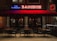 A cozy Thai restaurant with a neon sign in red and blue that reads 'Bangkok' and 'Thai Restaurant'. The exterior features a red awning over a sidewalk seating area with several tables and chairs. The tables are topped with small red candles, casting a warm glow. Inside, low lighting and curtains add to the intimate atmosphere.