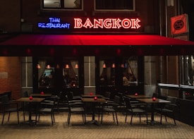 A cozy Thai restaurant with a neon sign in red and blue that reads 'Bangkok' and 'Thai Restaurant'. The exterior features a red awning over a sidewalk seating area with several tables and chairs. The tables are topped with small red candles, casting a warm glow. Inside, low lighting and curtains add to the intimate atmosphere.