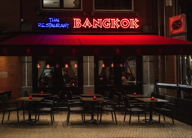A cozy Thai restaurant with a neon sign in red and blue that reads 'Bangkok' and 'Thai Restaurant'. The exterior features a red awning over a sidewalk seating area with several tables and chairs. The tables are topped with small red candles, casting a warm glow. Inside, low lighting and curtains add to the intimate atmosphere.
