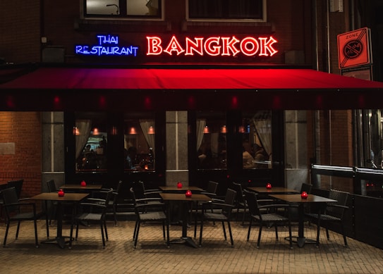 A cozy Thai restaurant with a neon sign in red and blue that reads 'Bangkok' and 'Thai Restaurant'. The exterior features a red awning over a sidewalk seating area with several tables and chairs. The tables are topped with small red candles, casting a warm glow. Inside, low lighting and curtains add to the intimate atmosphere.