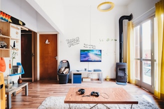 A cozy living room features a television mounted on the wall displaying a video game. The decor includes wooden furniture, a modern wood stove, and a light-colored shag rug. Bright yellow curtains allow natural light to fill the room. Shelves are adorned with books and decorative items. A modern coffee table has a pair of headphones and a game controller placed on it.