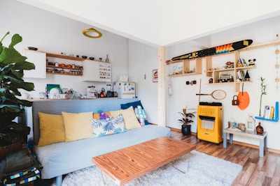 A cozy living room with a blue sofa adorned with colorful cushions featuring geometric and floral patterns. The walls are decorated with wooden shelves displaying books, small plants, and a variety of decorative items, including a toy car and a hanging paddle. On one side, a refrigerator is visible near a range of kitchenware, while a tall plant enhances the greenery in the space. A small wooden coffee table stands on a shaggy white rug, blending with the warm wooden flooring. The overall atmosphere is vibrant and eclectic, combining modern and rustic elements.