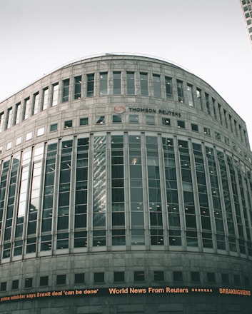 A large, modern office building with a curved facade and numerous windows. The building is labeled with the 'Thomson Reuters' logo. An electronic news ticker displays headlines along the bottom, contributing to the urban, corporate atmosphere.