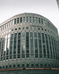 A large, modern office building with a curved facade and numerous windows. The building is labeled with the 'Thomson Reuters' logo. An electronic news ticker displays headlines along the bottom, contributing to the urban, corporate atmosphere.