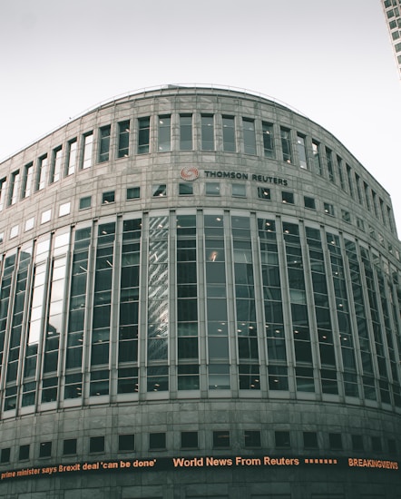 A large, modern office building with a curved facade and numerous windows. The building is labeled with the 'Thomson Reuters' logo. An electronic news ticker displays headlines along the bottom, contributing to the urban, corporate atmosphere.