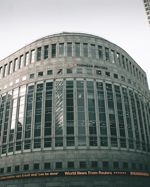 A large, modern office building with a curved facade and numerous windows. The building is labeled with the 'Thomson Reuters' logo. An electronic news ticker displays headlines along the bottom, contributing to the urban, corporate atmosphere.