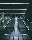 A modern escalator in a dimly lit indoor setting with sleek metallic railings and illuminated steps leading upward into a dark ceiling.