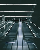A modern escalator in a dimly lit indoor setting with sleek metallic railings and illuminated steps leading upward into a dark ceiling.