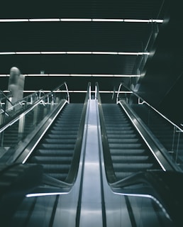 A modern escalator in a dimly lit indoor setting with sleek metallic railings and illuminated steps leading upward into a dark ceiling.