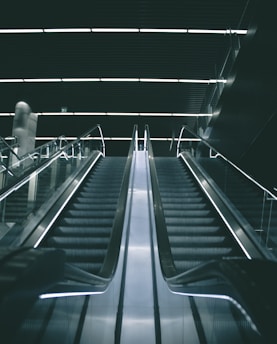 A modern escalator in a dimly lit indoor setting with sleek metallic railings and illuminated steps leading upward into a dark ceiling.