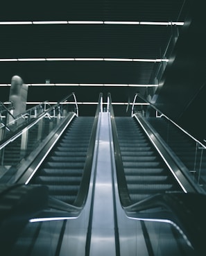 A modern escalator in a dimly lit indoor setting with sleek metallic railings and illuminated steps leading upward into a dark ceiling.