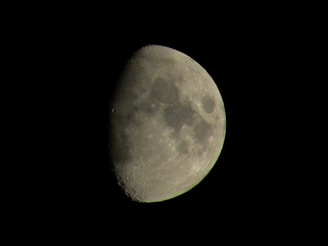 A detailed and clear view of the moon, capturing its rugged surface with visible craters and maria. The moon appears illuminated on one side, showing the distinct features of its surface against a stark black background.