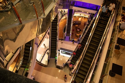 A multi-level shopping mall with several escalators connecting different floors. People are moving up and down the escalators, and some are walking on the floors around them. The interior features shiny surfaces, visible railings, and a mix of warm lighting with blue accents.