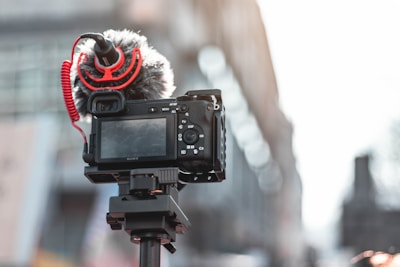 A digital camera is mounted on a tripod with a directional microphone attached. The microphone features a red coiled cable and a windscreen cover. The background is blurred, suggesting an urban environment.