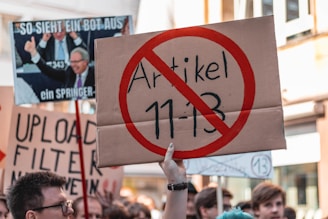 A protest scene with a crowd holding various signs, including one prominently displaying a sign with a red circle and line through 'Artikel 11-13'. Other signs in the background include messages about upload filters and internet freedom, conveyed in German. People appear engaged and concerned in an urban setting.