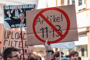 A protest scene with a crowd holding various signs, including one prominently displaying a sign with a red circle and line through 'Artikel 11-13'. Other signs in the background include messages about upload filters and internet freedom, conveyed in German. People appear engaged and concerned in an urban setting.
