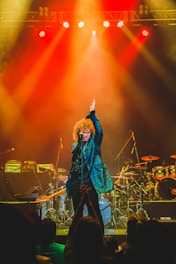 A performer with curly hair, wearing a shimmering blue jacket, stands on stage with one arm raised. The stage is equipped with musical instruments, including drums and guitars, and is illuminated by vibrant red and yellow stage lights. The audience can be seen in the foreground with some people clapping.