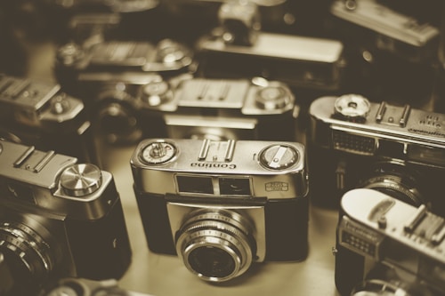 A collection of vintage cameras are closely arranged together. The cameras have a metallic finish with various dials, lenses, and nameplates visible. The image has a sepia tone, giving it a nostalgic and historic feel.