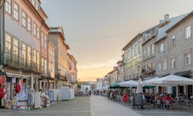 A charming European street with outdoor cafes lined with people enjoying their meals. The street is flanked by historic buildings adorned with large windows and traditional facades. Merchandise and colorful souvenirs are displayed outside shops on the left, while white and green awnings with brand logos mark the cafes on the right. The setting sun casts a warm glow across the scene, creating a peaceful atmosphere.