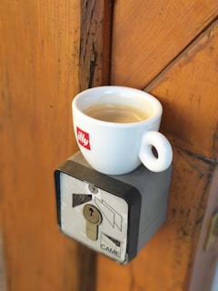 A white cup of coffee marked with the illy logo is placed atop a metal box containing a keyhole and the word CAME. The background consists of a wooden surface with visible grain and textures.