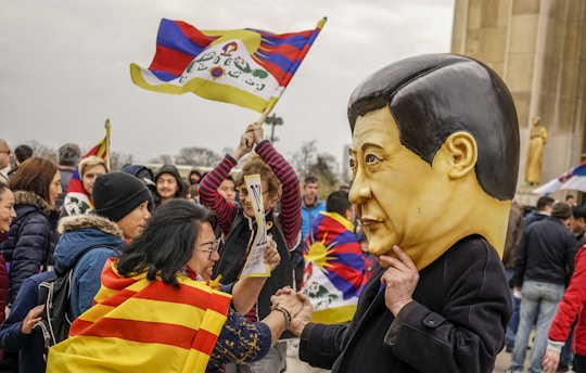 A group of people gathered in an outdoor setting, engaging in what appears to be a protest or demonstration. A person is wearing a large caricature mask, and others around them are holding flags with colorful designs, including a Tibetan flag. The atmosphere is vibrant with individuals interacting and expressing themselves actively.