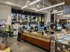 Inside a grocery store, several self-checkout counters are aligned with people actively scanning items. Shoppers are engaged in processing their purchases amidst a modern, well-lit interior. There are visible retail displays and magazines near the exits.