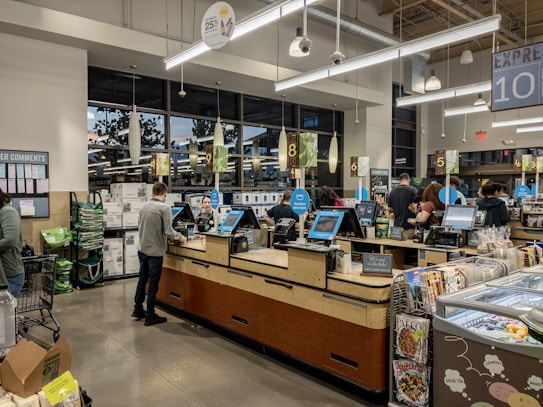 Inside a grocery store, several self-checkout counters are aligned with people actively scanning items. Shoppers are engaged in processing their purchases amidst a modern, well-lit interior. There are visible retail displays and magazines near the exits.