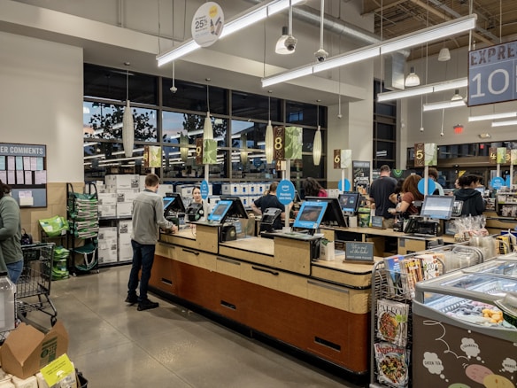 Inside a grocery store, several self-checkout counters are aligned with people actively scanning items. Shoppers are engaged in processing their purchases amidst a modern, well-lit interior. There are visible retail displays and magazines near the exits.