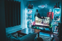 A room filled with collectible action figures and memorabilia, including figures from popular franchises like Star Wars. Illuminated glass display cabinets line the walls, each showcasing various figurines. A television displays a nature scene, sitting atop a stand filled with assorted gaming consoles or entertainment devices. The room has a modern and organized appearance with a cozy ambiance. A zebra-patterned rug lies under a wooden coffee table, adding a touch of style.
