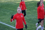 A group of people wearing matching red athletic jackets and black gloves are on a grassy sports field. One person in the foreground is smiling and making a peace sign with their hand. Others in the background seem to be walking or preparing for an activity.