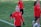 A group of people wearing matching red athletic jackets and black gloves are on a grassy sports field. One person in the foreground is smiling and making a peace sign with their hand. Others in the background seem to be walking or preparing for an activity.