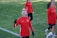 A group of people wearing matching red athletic jackets and black gloves are on a grassy sports field. One person in the foreground is smiling and making a peace sign with their hand. Others in the background seem to be walking or preparing for an activity.