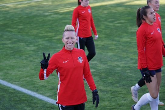 A group of people wearing matching red athletic jackets and black gloves are on a grassy sports field. One person in the foreground is smiling and making a peace sign with their hand. Others in the background seem to be walking or preparing for an activity.