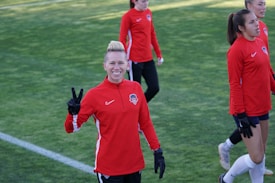 A group of people wearing matching red athletic jackets and black gloves are on a grassy sports field. One person in the foreground is smiling and making a peace sign with their hand. Others in the background seem to be walking or preparing for an activity.