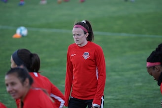 A group of athletes wearing red sports jerseys on a grassy field, one person is more prominent in the foreground, looking focused. The jerseys have a sports logo on the chest, and some players are wearing pink headbands. In the background, a soccer ball is visible.