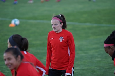 A group of athletes wearing red sports jerseys on a grassy field, one person is more prominent in the foreground, looking focused. The jerseys have a sports logo on the chest, and some players are wearing pink headbands. In the background, a soccer ball is visible.