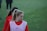 A person with a ponytail wearing a red sports jacket and a white training bib stands on a grassy field. The individual appears to be focused or concentrating, with others in similar attire visible in the background.