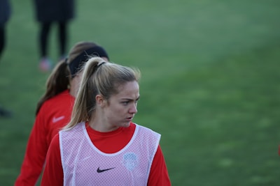 A person with a ponytail wearing a red sports jacket and a white training bib stands on a grassy field. The individual appears to be focused or concentrating, with others in similar attire visible in the background.